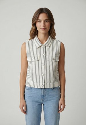 Giovane donna con gilet bianco senza maniche a bottoni con strisce verticali nere e jeans azzurri a vita alta, in piedi davanti a uno sfondo neutro.