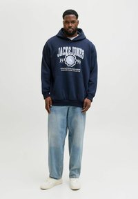 Marineblauwe hoodie met witte grafische tekst en logo op de voorkant, ontspannen pasvorm, gecombineerd met lichtblauwe jeans en witte sneakers. Glad textuur.