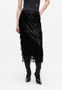 Jupe à franges en sequins noirs avec une coupe mi-longue. Elle possède une taille lisse et se marie parfaitement avec un pantalon noir près du corps et des bottines noires brillantes.