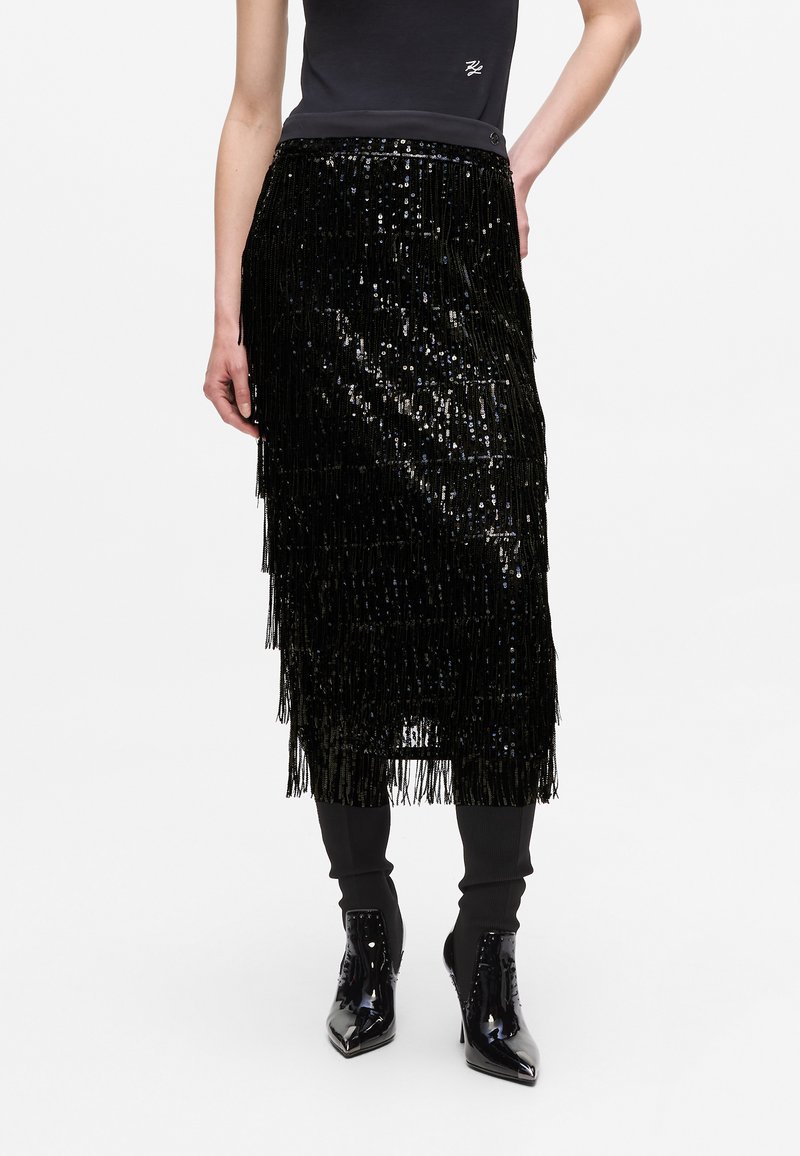 Jupe à franges en sequins noirs avec une coupe mi-longue. Elle possède une taille lisse et se marie parfaitement avec un pantalon noir près du corps et des bottines noires brillantes.
