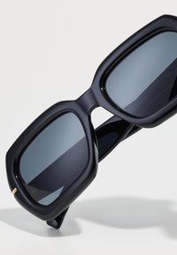 Crne velike sunčane naočale s debelim okvirom, ravnim staklima i sjajnim završetkom. Imaju mali zlatni detalj na okviru.