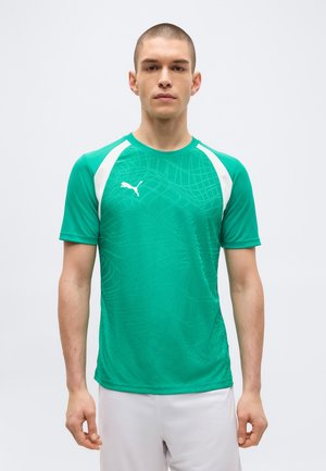 Mann trägt ein grünes Puma-Sportshirt mit weißen Akzenten und dezentem Muster, steht vor einem schlichten hellgrauen Hintergrund.