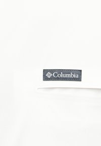 Ετικέτα της μάρκας Columbia σε άσπρο ύφασμα πάνω από μια οριζόντια τσέπη με διακριτική σκίαση.