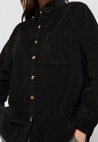 Chemise en velours côtelé noire à manches longues, devant boutonné, col, poche poitrine simple et boutons dorés pour le contraste. Texture douce.