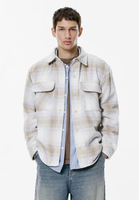 Plaid shirt in light tones of beige and blue, with two chest pockets, button closure, and a relaxed fit, layered over a brown base.