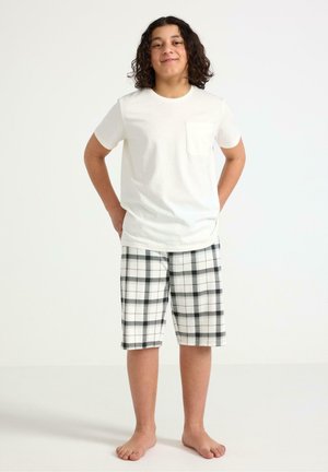 Nuori henkilö, jolla on kiharat tummat hiukset, seisoo paljain jaloin, pukeutuneena valkoiseen lyhythihaiseen paitaan ja mustavalkoisiin ruudullisiin shortseihin yksiväristä taustaa vasten.