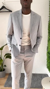 Világosszürke, csíkos öltöny texturált anyagból, egysoros kabáttal, amelynek két gombja van, és keskeny nadrággal, fehér ing fölött viselve.