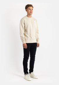 Beige sweatshirt with long sleeves and a crew neckline, paired with black slim-fit jeans and white sneakers. Features a small logo.