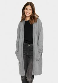 Cardigan lungo grigio, leggero, con davanti aperto, maniche lunghe e tasche laterali, abbinato a un top nero e jeans scuri.