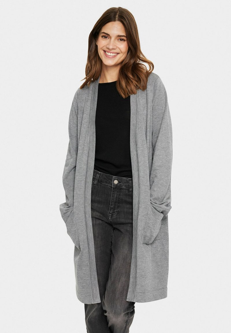 Cardigan lungo grigio, leggero, con davanti aperto, maniche lunghe e tasche laterali, abbinato a un top nero e jeans scuri.