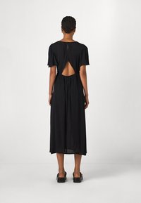 Robe noire mi-longue avec des manches courtes, détail de dos ouvert, taille cintrée et tissu fluide, associée à des chaussures à talons carrés noires.