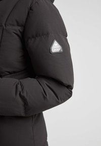Chaqueta aislante negra con textura acolchada, con un parche de logo triangular plateado en la manga izquierda. Superficie de tela suave.