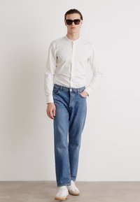 White button-up shirt with a band collar, paired with blue jeans and white sneakers. Model wears dark sunglasses.