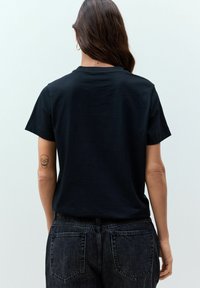 T-shirt nera a girocollo con maniche corte, realizzata in morbido tessuto. Il modello indossa jeans scuri. Un tatuaggio di teschio è visibile sul braccio sinistro.