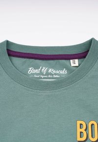 Minzgrünes T-Shirt mit kurzen Ärmeln, lila Innenkragen und großem gelben "BO"-Grafik. Hergestellt aus Bio-Baumwolle, mit glatter Textur.