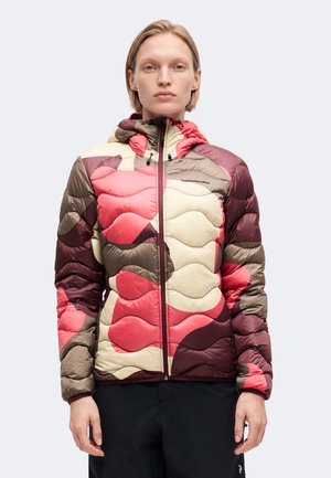 Personne portant une veste matelassée à capuche avec des patchs abstraits rouges, beige et marron, debout face à un fond clair.