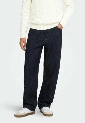 Personne portant un jean ample bleu foncé, un pull à torsades blanc et des baskets blanches avec des détails en suède beige.