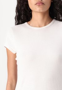 T-shirt en coton blanc avec des manches courtes roulées, col rond et une texture lisse. Présente un design minimaliste et une coupe ajustée.