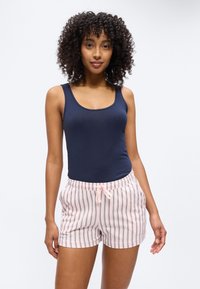 Γυναίκα με σγουρά μαλλιά φοράει μπλε σκούρο αμάνικο τοπ και ροζ και λευκές ριγέ βερμούδες με κορδόνι, στέκεται μπροστά σε ένα απλό φόντο.