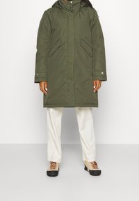 Olivgrön parka med dragkedja och tryckknappar, två framfickor, huva och justerbara ärmar. Bärs över ljusa byxor och skor.