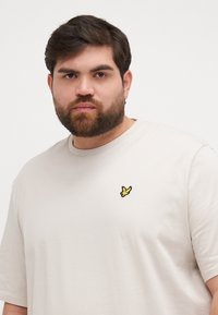 T-shirt beige clair à manches courtes avec un col rond et un petit logo d'oiseau jaune brodé sur le côté gauche de la poitrine. Texture lisse.