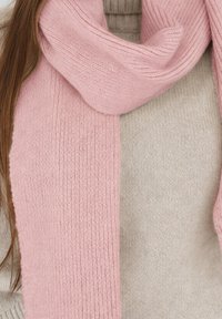 Écharpe en tricot rose avec une texture côtelée, enroulée autour d'un pull beige. L'écharpe présente une finition douce et lisse.