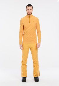 Maglione arancione in pile a maniche lunghe con colletto a mezza zip, abbinato a pantaloni da neve gialli. Stivali da neve neri completano l'outfit.