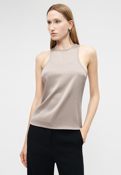 Femme aux cheveux blonds raides portant des boucles d'oreilles créoles, un débardeur taupe sans manches à col montant, et un pantalon noir, debout avec une main dans la poche.