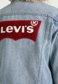 Světlá modrá džínová bunda s velkou červenobílou nášivkou s logem Levi's z froté na zádech, s klasickým límcem a prošitými detaily.