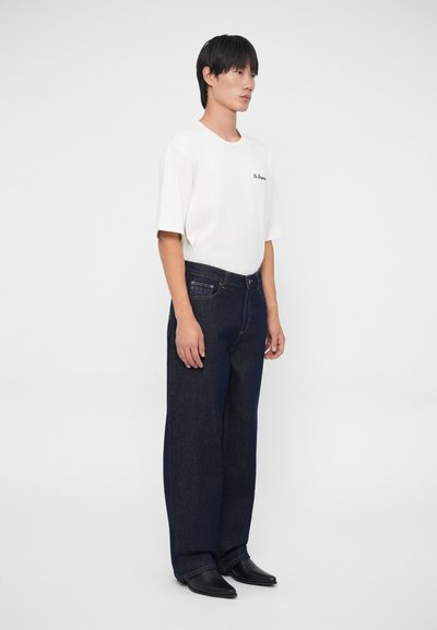 Camiseta blanca de manga corta con bordado negro combinada con jeans de mezclilla de pierna ancha en azul oscuro y botas de cuero negras. Fondo liso.