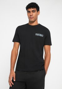 Sort bomulds T-shirt med korte ærmer, rund halsudskæring og et lille hvidt logo på venstre bryst. Glat tekstur, normal pasform.