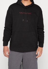 Persona vestida con sudadera negra con el texto rojo "BASECAMP Sigue el Sendero", pantalones grises, de pie contra un fondo liso.