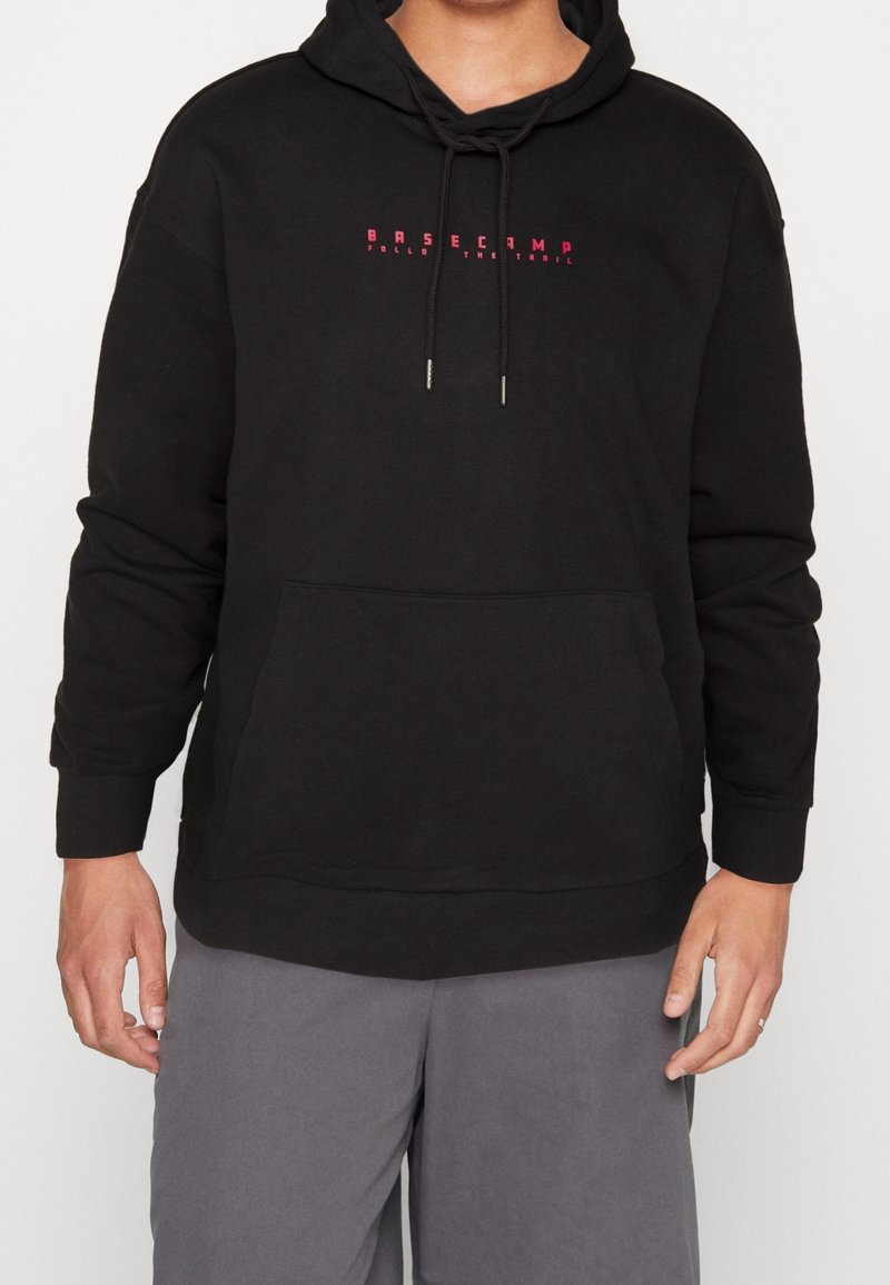 Persona vestida con sudadera negra con el texto rojo "BASECAMP Sigue el Sendero", pantalones grises, de pie contra un fondo liso.