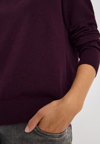 Bordowy sweter z dzianiny z prążkowanymi mankietami i dołem, z luźnym krojem i kieszenią po bokach w szarych dżinsach. Widoczna miękka tekstura.