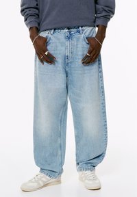 Persoon in losse lichtblauwe jeans, witte sneakers, een grijze longsleeve top, en zilveren ringen en armbanden aan beide handen.