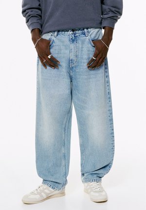 Person trägt lockere hellblaue Jeans, weiße Sneakers, ein langärmeliges graues Oberteil sowie silberne Ringe und Armbänder an beiden Händen.