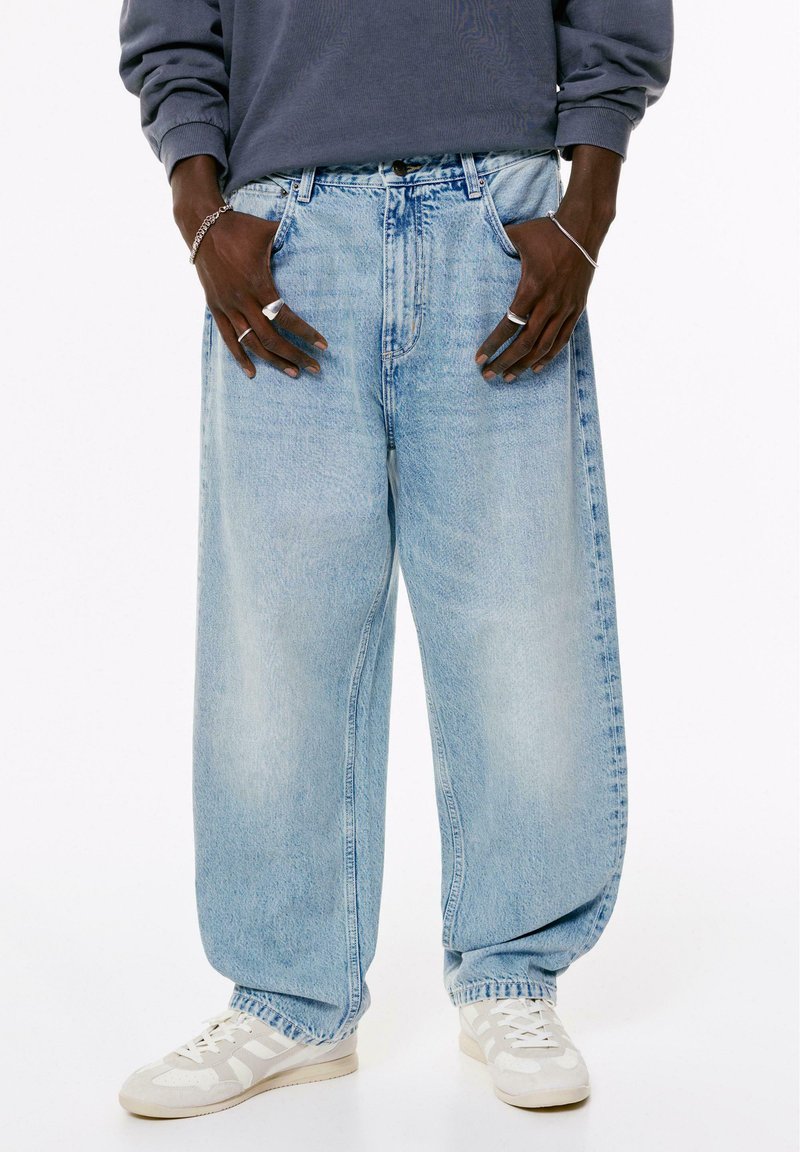 Persoon in losse lichtblauwe jeans, witte sneakers, een grijze longsleeve top, en zilveren ringen en armbanden aan beide handen.