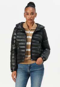 Zwarte pufferjack met een glanzende afwerking, voorzien van een capuchon, ritszakken en geribde manchetten, gedragen boven een beige gestreepte trui en blauwe jeans.
