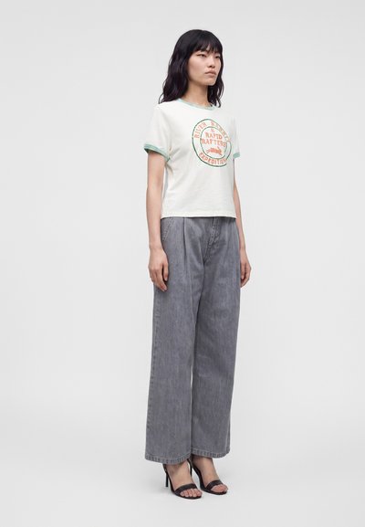 Jeune femme aux cheveux noirs portant un t-shirt blanc avec un motif graphique, un pantalon gris ample et des sandales noires à talons hauts, debout devant un fond uni.