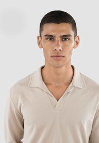 Hellbeige Pullover mit V-Ausschnitt und Kragen. Der Stoff hat eine strukturierte Oberfläche und die Passform ist körpernah, mit langen Ärmeln bis zu den Handgelenken.