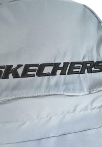 Chaqueta acolchada de un gris claro, con un destacado logotipo bordado en negro de "SKECHERS". Tela suave con un ligero brillo y detalles de costura.