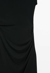 Détail d'une robe noire ajustée montrant un tissu lisse avec un léger drapé froncé sur le côté droit et une couture horizontale à la taille.