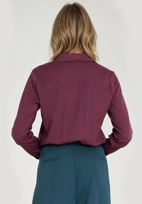 Burgundowa koszula z długim rękawem i kołnierzykiem oraz szwem z tyłu, w zestawieniu z ciemnozielonymi spodniami. Gładka tekstura materiału o dopasowanym kroju.