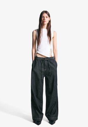 White sleeveless top with side button detail, paired with loose dark denim pants featuring an elastic waistband and drawstring.