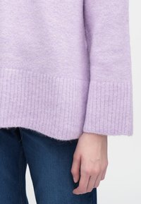 Ohlapen pulover iz sivke v mehki pletenini, s rebrastimi manšetami in robom ter rahlo ukrivljenim spodnjim robom, kombiniran z temno modrimi kavbojkami.