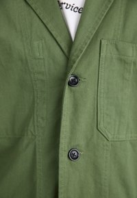 Olivengrønn blazer med notch krage, svarte knapper og en lomme med klaff. Laget av strukturert stoff med synlige sømdetaljer.