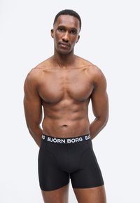 Μυώδης άνδρας που στέκεται με τα χέρια πίσω από την πλάτη, φοράει μαύρα μποξέρ Björn Borg σε απλό φωτεινό φόντο.