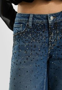 Spijkerbroek met een getailleerd ontwerp, voorzien van donkerblauwe stof, verweerde details en zwarte en zilveren decoratieve studs aan de voorkant.