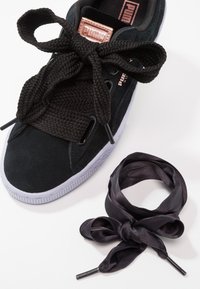 Baskets Puma en daim noir avec de gros lacets noirs noués en nœud et une paire supplémentaire de lacets fins, brillants et noirs enroulés à côté.