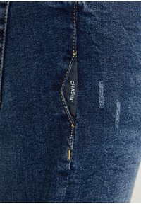 Jean en denim bleu foncé avec une texture déchirée, comportant une poche cachée avec une étiquette noire inscrite "CHASIN." et des surpiqûres jaunes contrastantes.